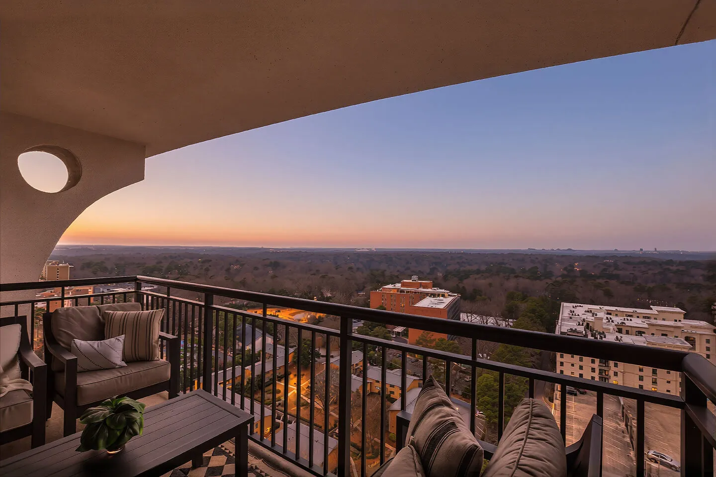 High-rise balcony with outdoor seating overlooking a tree-lined cityscape at sunset